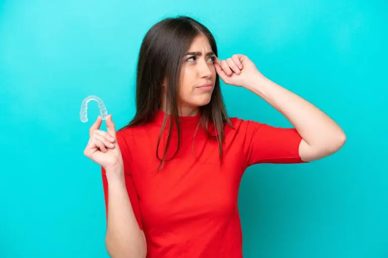 A woman in a red shirt, standing against a turquoise background, holds a clear dental aligner and rubs one eye—illustrating clear aligner treatments offered by Andros Orthodontics in Pasco, WA.