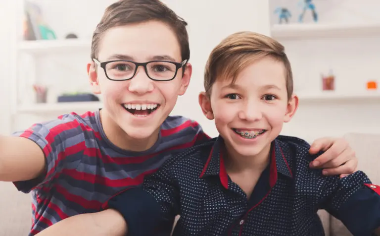 On a couch in Pasco, WA, two smiling boys sit close together—one in glasses and a striped shirt, the other sporting Incognito Braces and a dark patterned shirt—showcasing happy patients of Andros Orthodontics.