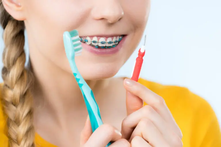 At Andros Orthodontics in Pasco, WA, a person in a yellow shirt smiles with clean braces, holding a toothbrush in one hand and an interdental brush in the other.