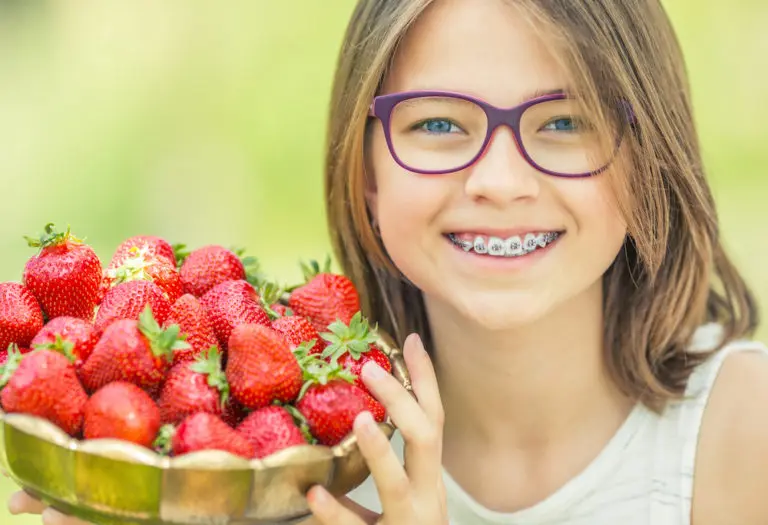 A smiling child with braces and purple glasses enjoys a bowl of fresh strawberries—a perfect braces-friendly snack—outside, as seen at Andros Orthodontics in Pasco, WA.