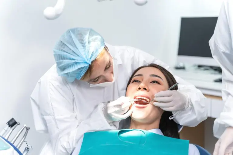 At Andros Orthodontics in Pasco, WA, a dentist wearing protective clothing examines a patient's teeth using dental tools while the patient sits with a bib and mouth open.