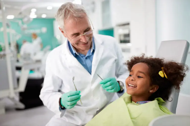At Andros Orthodontics in Pasco, WA, a dentist in a white coat and gloves warmly smiles at a young girl during her first visit. The girl, wearing a green bib and holding dental tools, returns the smile while seated in the dental chair.