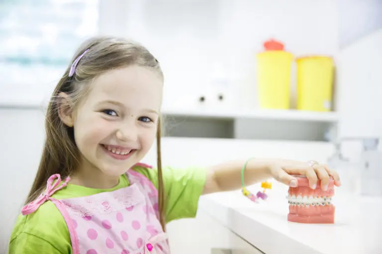 A young girl excitedly smiles and holds a braces dental model during her first visit at Andros Orthodontics in Pasco, WA.