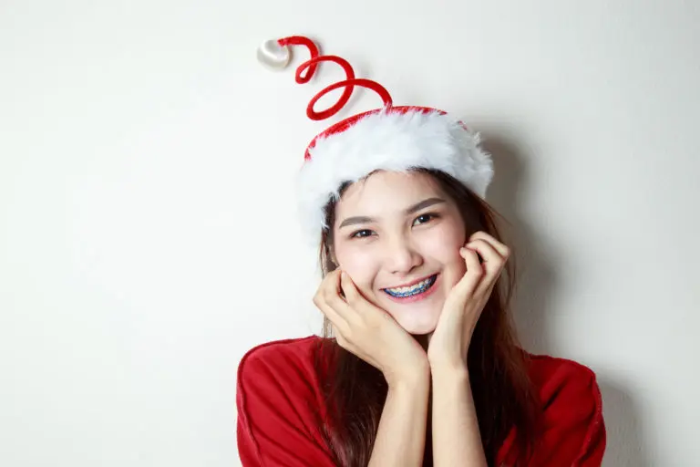 A young woman with long hair and blue braces, wearing a red and white Santa hat with a spiral top, smiles joyfully for Andros Orthodontics in Pasco, WA, capturing the festive holiday spirit against a plain white wall.