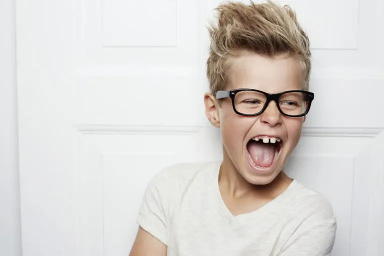 A young boy with short, tousled blonde hair and black glasses laughs, showing a tooth gap, while standing in front of a white paneled door—capturing the kind of smiles created by Andros Orthodontics in Pasco, WA.