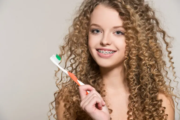 A young woman with curly hair and braces smiles at the camera while holding a toothbrush and toothpaste, representing Andros Orthodontics in Pasco, WA.
