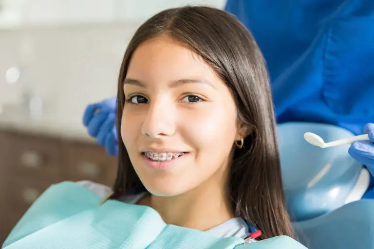At Andros Orthodontics in Pasco, WA, a teenage girl with braces sits in a dental chair wearing a bib as a dental professional in blue scrubs and gloves stands nearby holding a dental tool.