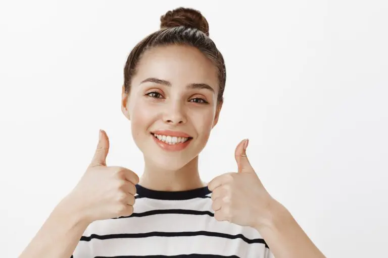 A young woman in a striped shirt smiles and gives two thumbs up in front of a plain white background, showcasing her happiness with Andros Orthodontics in Pasco, WA.