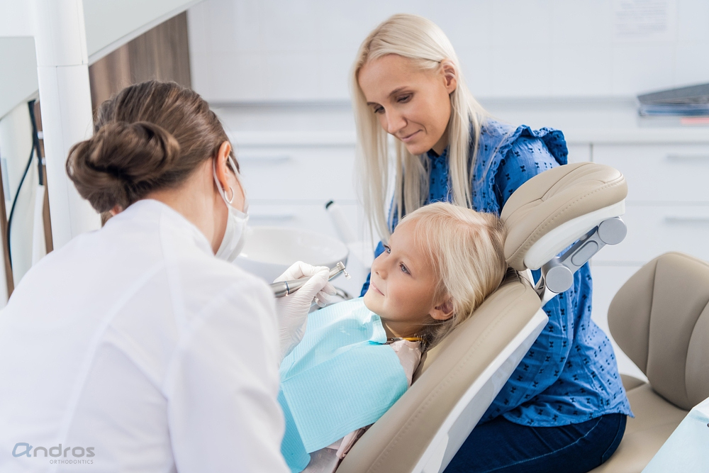 A young child sits in a dental chair receiving treatment from a dentist while an adult woman, likely a parent, sits nearby watching after being explained about how to fix overbite in Pasco, WA.