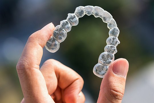 A clear Invisalign dental aligner, used for orthodontic teeth straightening, is displayed in a hand against a blurred background at Andros Orthodontics in Pasco, WA.