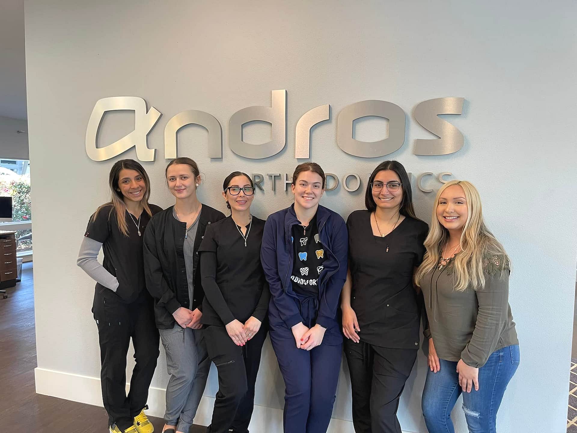 Six women from the team at Andros Orthodontics in Pasco, WA smile together in front of a wall sign, all dressed in work attire.