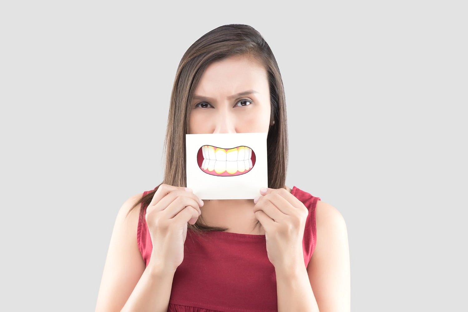 A woman in a red sleeveless top stands against a plain light background, holding a paper with a cartoon smile of white teeth over her mouth to conceal her underbite—an image representing Andros Orthodontics in Pasco, WA.