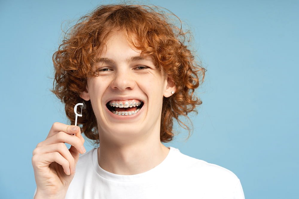 A teen with curly hair and braces smiles while holding a dental floss pick against a blue background, emphasizing the importance of proper dental hygiene for both teens and Adult Orthodontics patients at Andros Orthodontics in Pasco, WA.