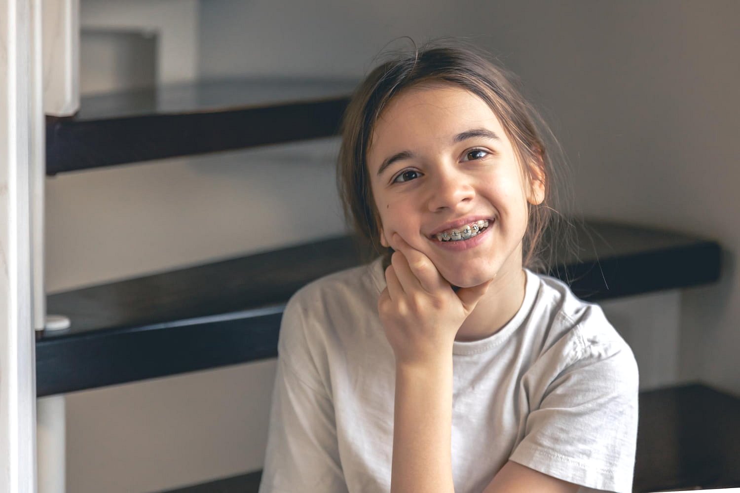 A young person with long hair wearing a light-colored t-shirt and traditional braces, seen smiling with one hand on their chin while sitting on stairs, represents the confident results from Andros Orthodontics — a trusted Children’s Orthodontist in Pasco, WA.