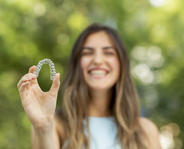 A woman smiles while holding a clear dental aligner, set against a blurred green outdoor background, considering if Invisalign is covered by insurance at Andros Orthodontics in Pasco, WA.