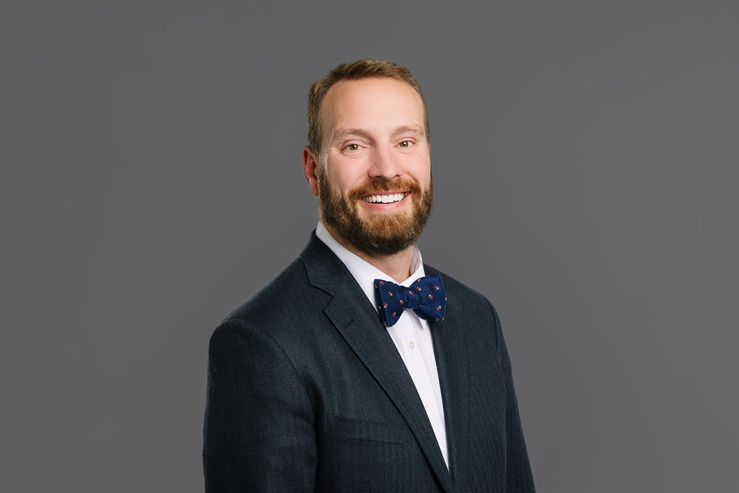 Dr. Nicholas Andros is pictured smiling at the camera in a dark suit with a white shirt and navy blue bow tie against a plain gray background. Dr. Nicholas Andros is orthodontics expert at Andros Orthodontics in Pasco, WA.