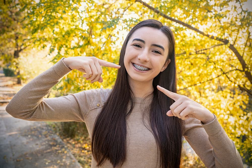 A young woman with long dark hair stands outdoors near yellow-leaved trees, smiling and pointing to her metal braces—showcasing her Andros Orthodontics treatment in Pasco, WA.