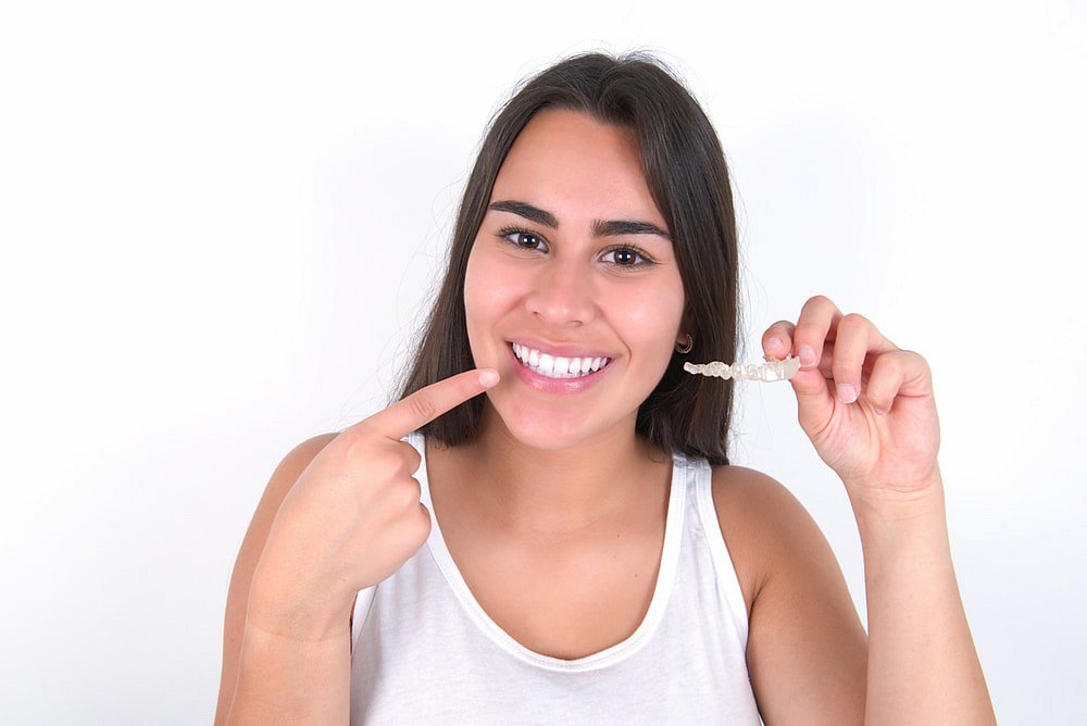 A woman smiles and points to her teeth while holding a clear aligner, showcasing the benefits of an Invisalign Payment Plan at Andros Orthodontics in Pasco, WA.