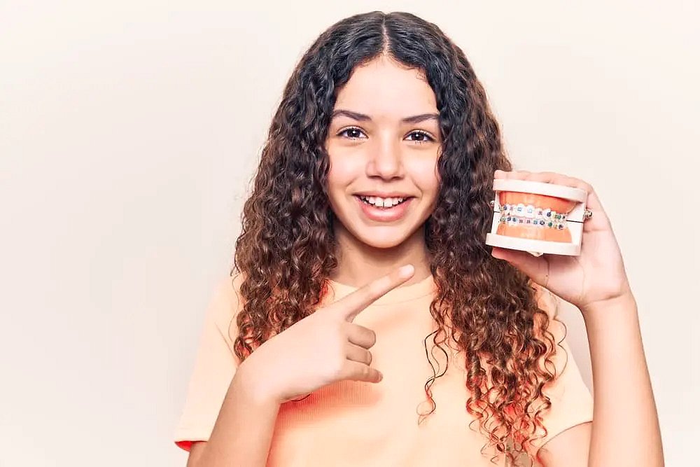 Andros Orthodontics A girl with curly hair smiles and points to a dental model with braces at Andros Orthodontics in Pasco, WA, showcasing orthodontic treatment options and raising awareness about Braces Cost and affordability.
