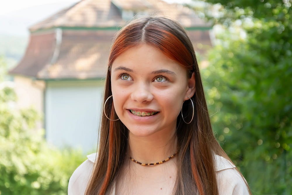 A teen girl with straight brown hair, green eyes, and metal braces—fitted by Andros Orthodontics in Pasco, WA—stands outdoors wearing hoop earrings and a bead necklace, with greenery and a house in the background.