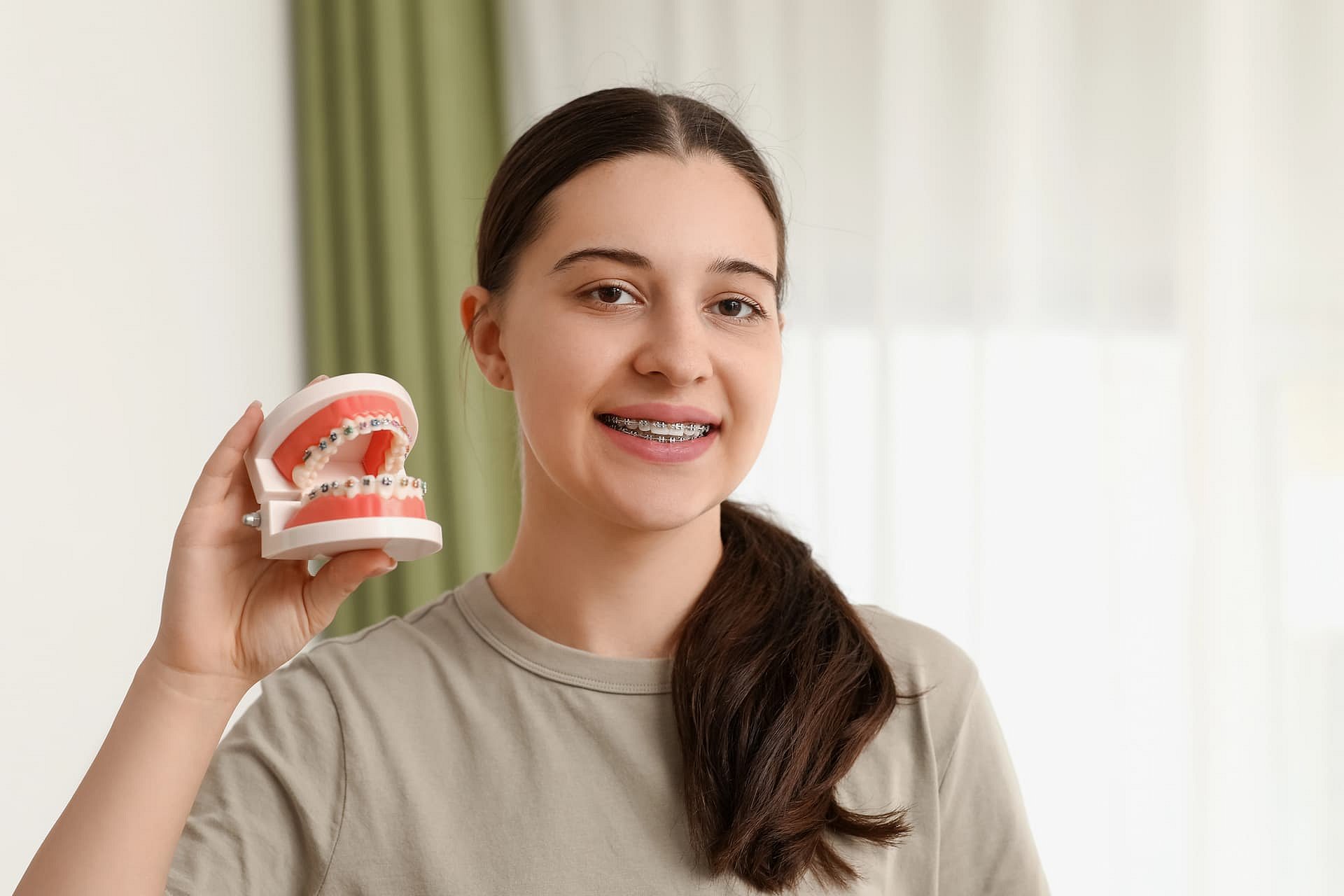A young woman with metal braces stands near a window with a green curtain, smiling and holding a dental model with braces—representing Andros Orthodontics in Pasco, WA.