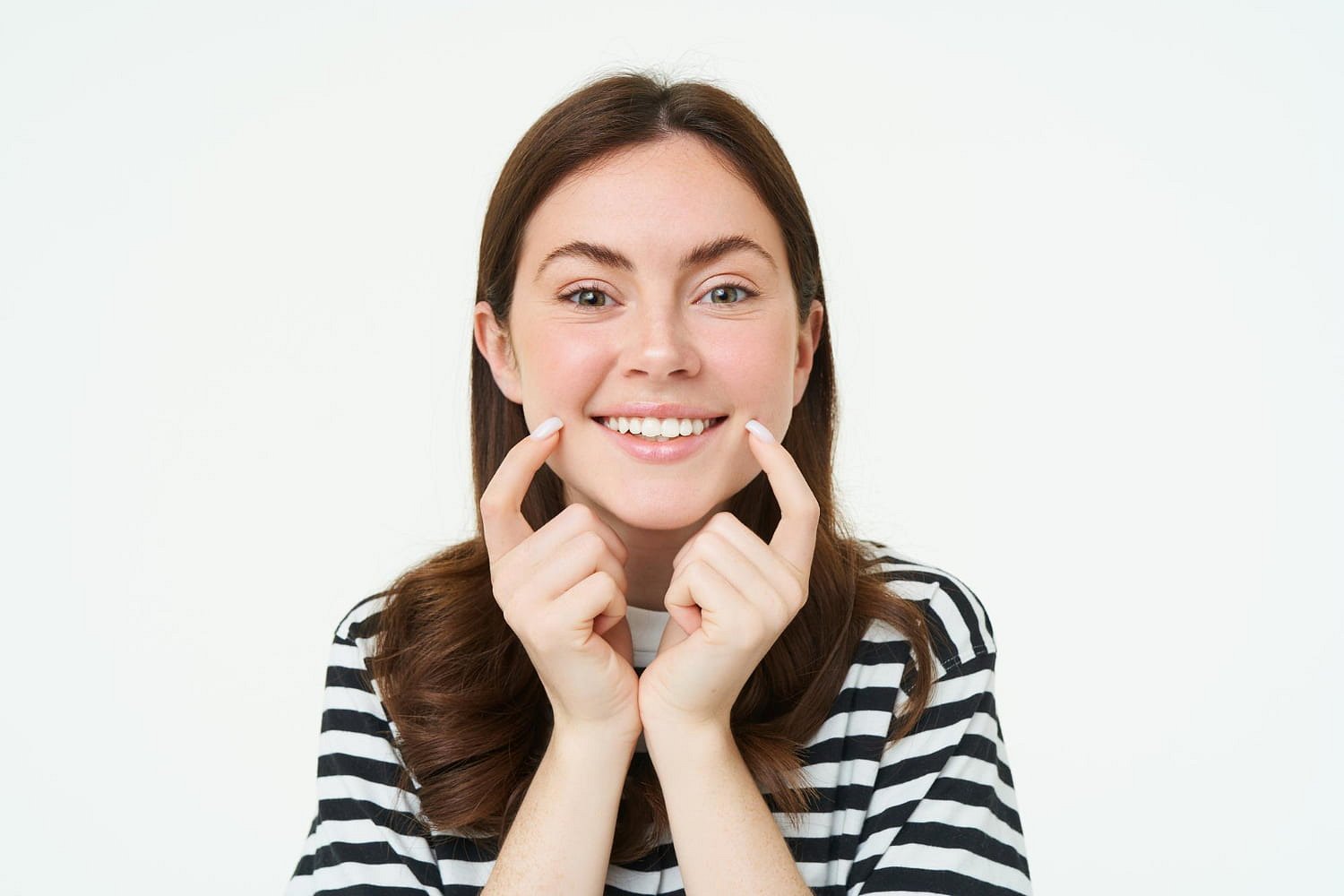 A woman with long brown hair and a black-and-white striped shirt smiles and points to her teeth, highlighting her overbite, in a photo for Andros Orthodontics in Pasco, WA.