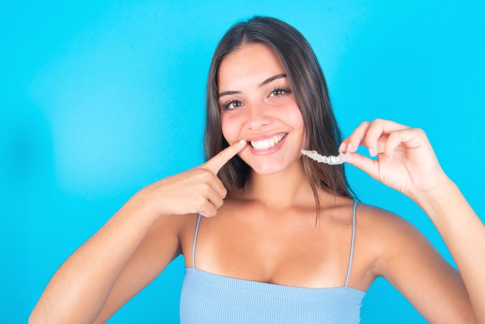 A woman in a blue tank top smiles and points at her teeth while holding a clear dental aligner, showcasing how Invisalign may be covered by insurance at Andros Orthodontics in Pasco, WA, against a bright blue background.