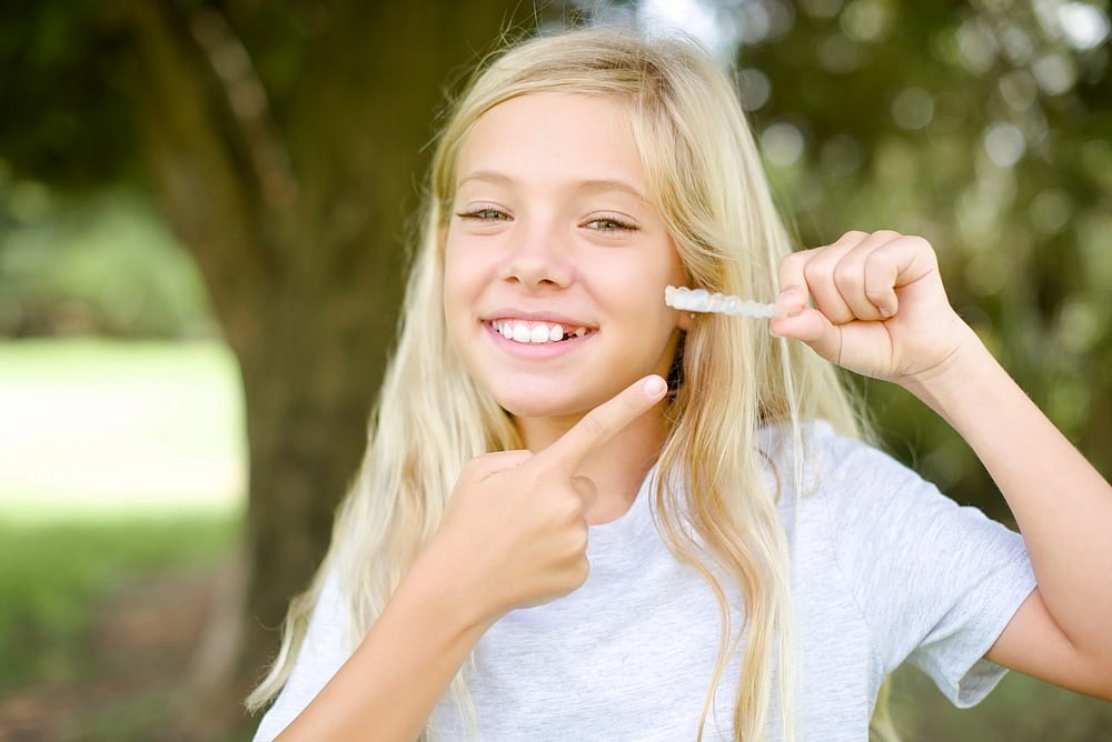 A smiling girl with long blonde hair in a gray t-shirt stands outdoors, holding up and pointing to Clear Aligners—available from Andros Orthodontics, a leading Children’s Orthodontist in Pasco, WA.
