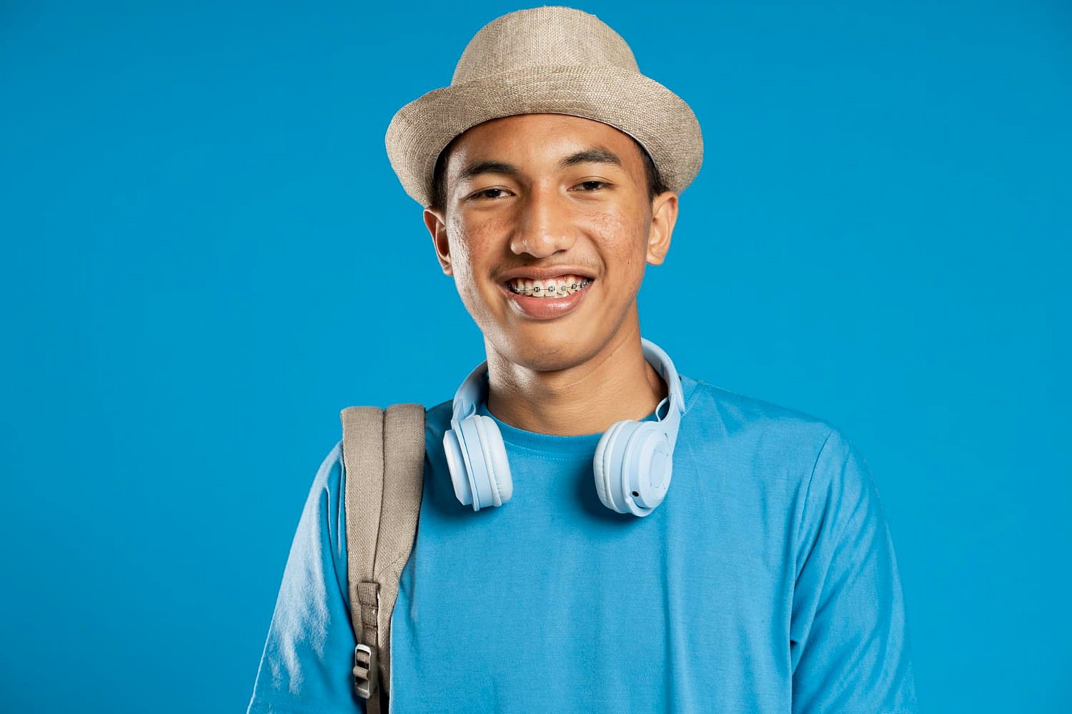 Smiling confidently against a blue background, a young person with metal braces—seen wearing a hat, blue shirt, headphones around their neck, and a backpack—perfectly captures the spirit of Andros Orthodontics in Pasco, WA.