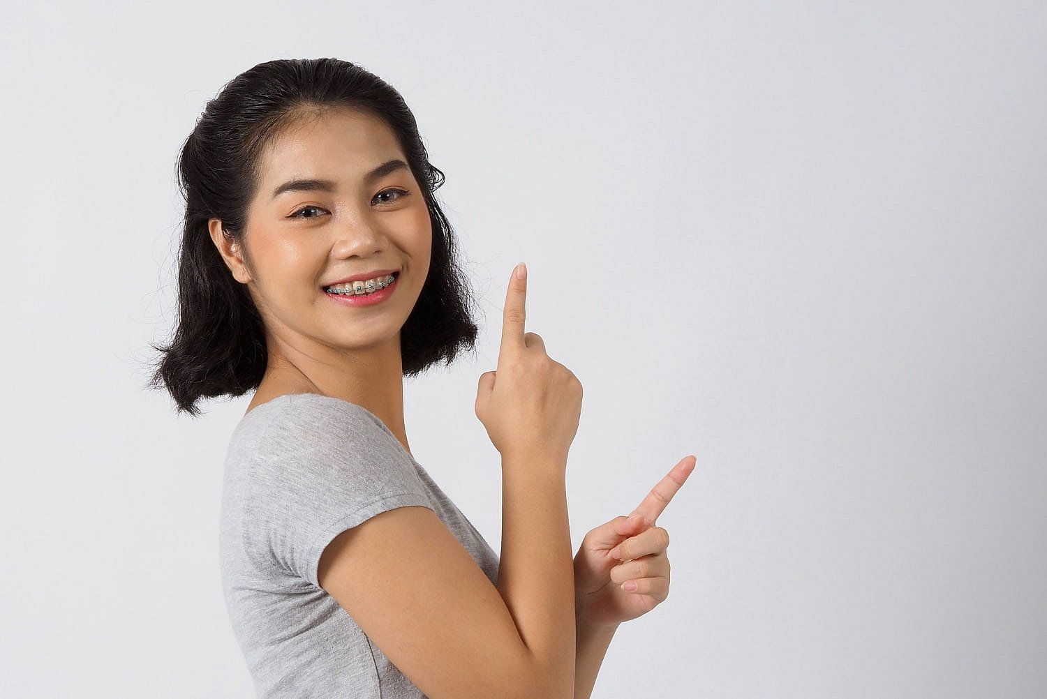 A young woman with short black hair and braces, displaying a slight underbite, smiles and gestures upward and to the side against a plain white background—an image representing Andros Orthodontics in Pasco, WA.