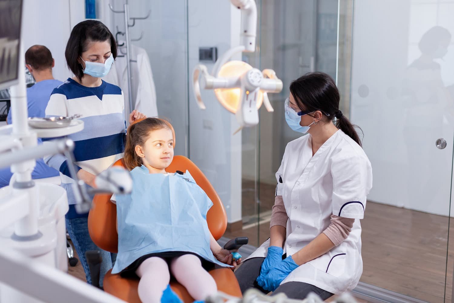 At Andros Orthodontics in Pasco, WA, a child sits in a modern children's orthodontist clinic, wearing a protective bib and accompanied by an adult and a mask-wearing dentist.