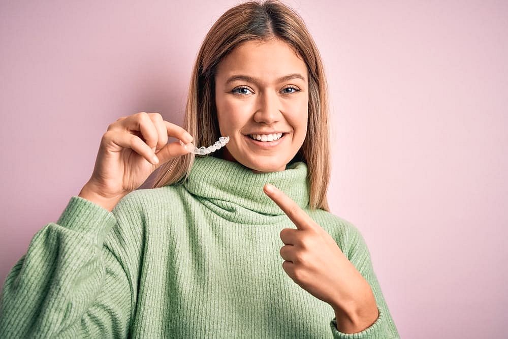 At Andros Orthodontics in Pasco, WA, a young woman in a green sweater smiles and points at Clear Aligners she holds against a light pink background.