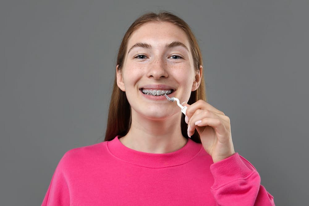 Andros Orthodontics A young woman in a pink sweatshirt uses a dental floss pick while considering braces cost at Andros Orthodontics in Pasco, WA, with braces visible against a plain gray background.