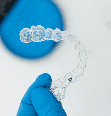 A gloved hand displays a clear dental aligner, highlighting the discreet Invisalign design at Andros Orthodontics in Pasco, WA. A blue circular object in the corner suggests convenient options like the Invisalign Payment Plan against a clean white background.