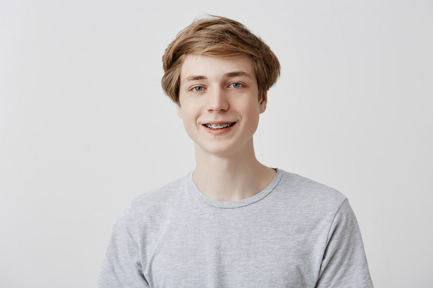 A teenage boy with light brown hair and braces confidently smiles at the camera in a grey t-shirt against a plain white background, showcasing results from Andros Orthodontics in Pasco, WA.