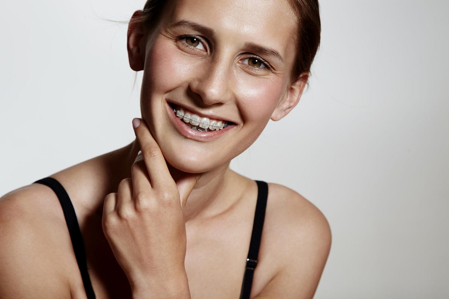 A young woman in a black strap top smiles at the camera, showing off her clear braces and gently touching her chin. This image captures the confidence that Adult Orthodontics can bring to patients at Andros Orthodontics in Pasco, WA.