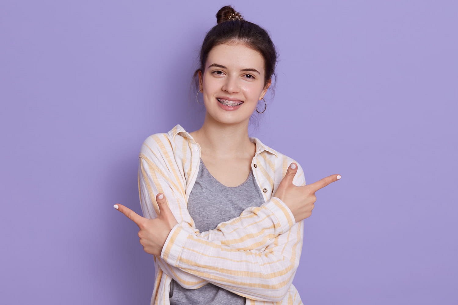 A young woman with metal braces, her hair in a bun, and dressed in a striped shirt over a gray t-shirt stands against a purple background, smiling and pointing both index fingers sideways. This image represents Andros Orthodontics in Pasco, WA.