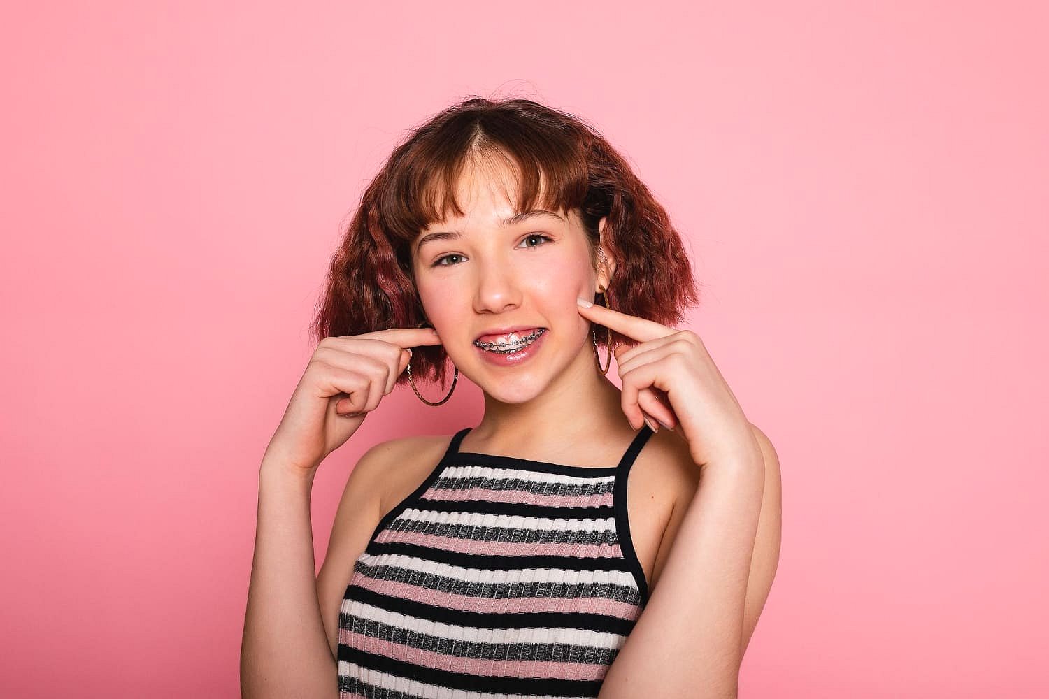 Showcasing her choice in the Invisalign vs Braces debate, a teenager with braces and short brown hair stands against a pink background, smiling and pointing to her cheeks while wearing a striped sleeveless top—representing Andros Orthodontics in Pasco, WA.