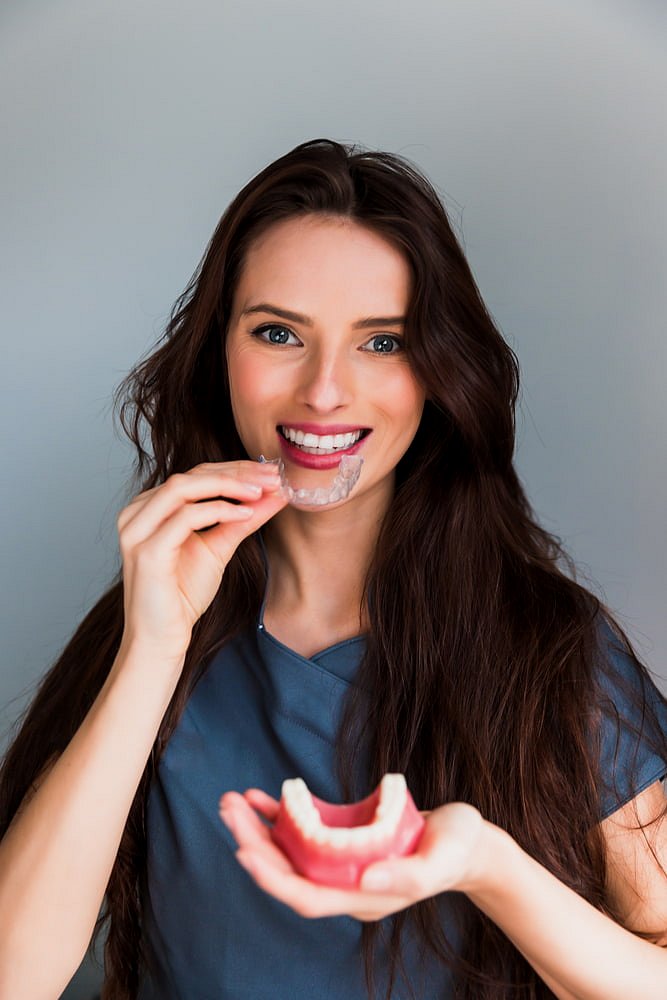 A woman with long brown hair, smiling at the camera, holds a dental model and places Invisalign® Clear Aligners on her teeth—highlighting Adult Orthodontics at Andros Orthodontics in Pasco, WA.