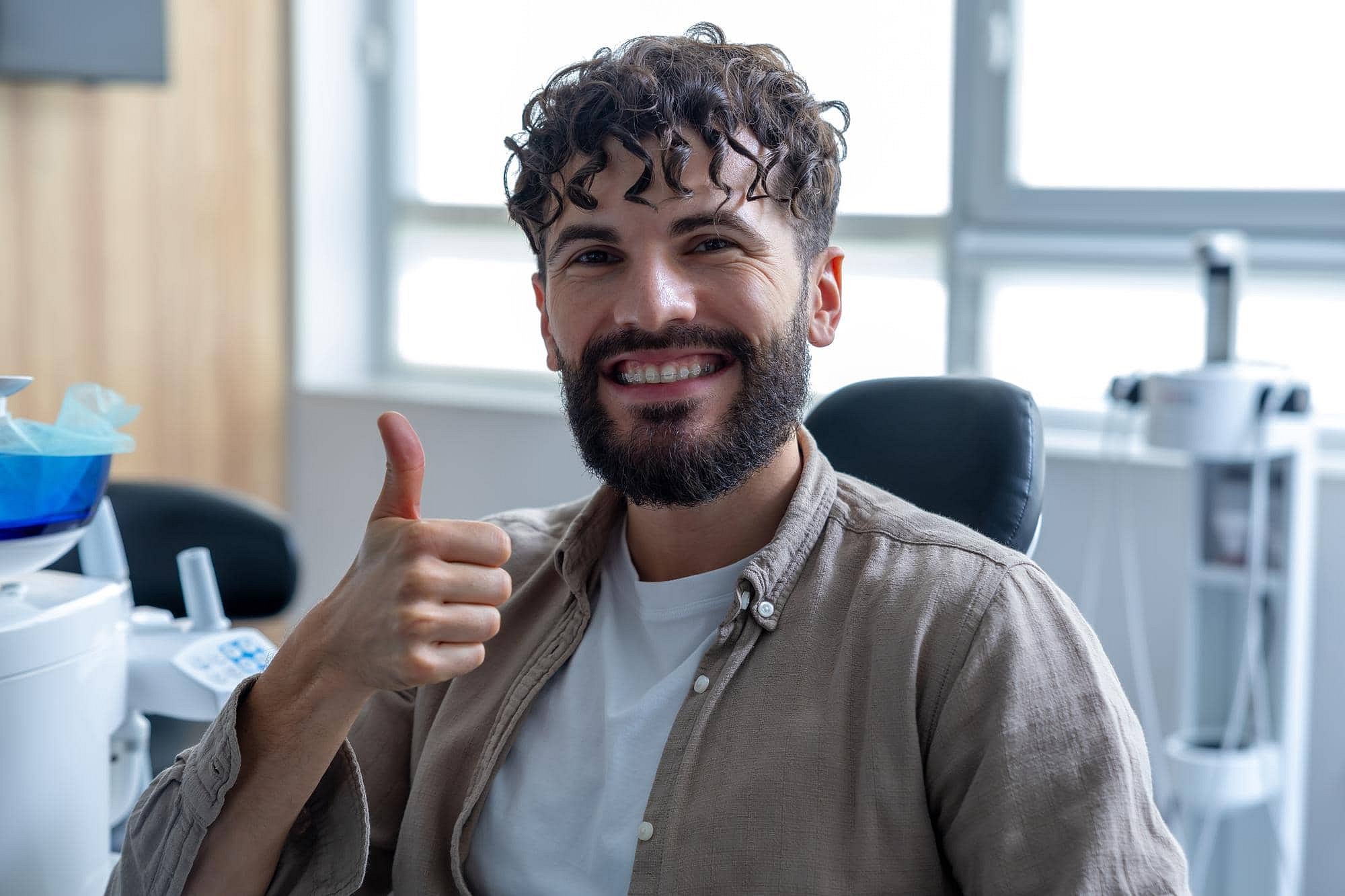 At Andros Orthodontics in Pasco, WA, a man with curly hair and a beard smiles and gives a thumbs up in the dental office, proudly displaying his new clear ceramic braces.