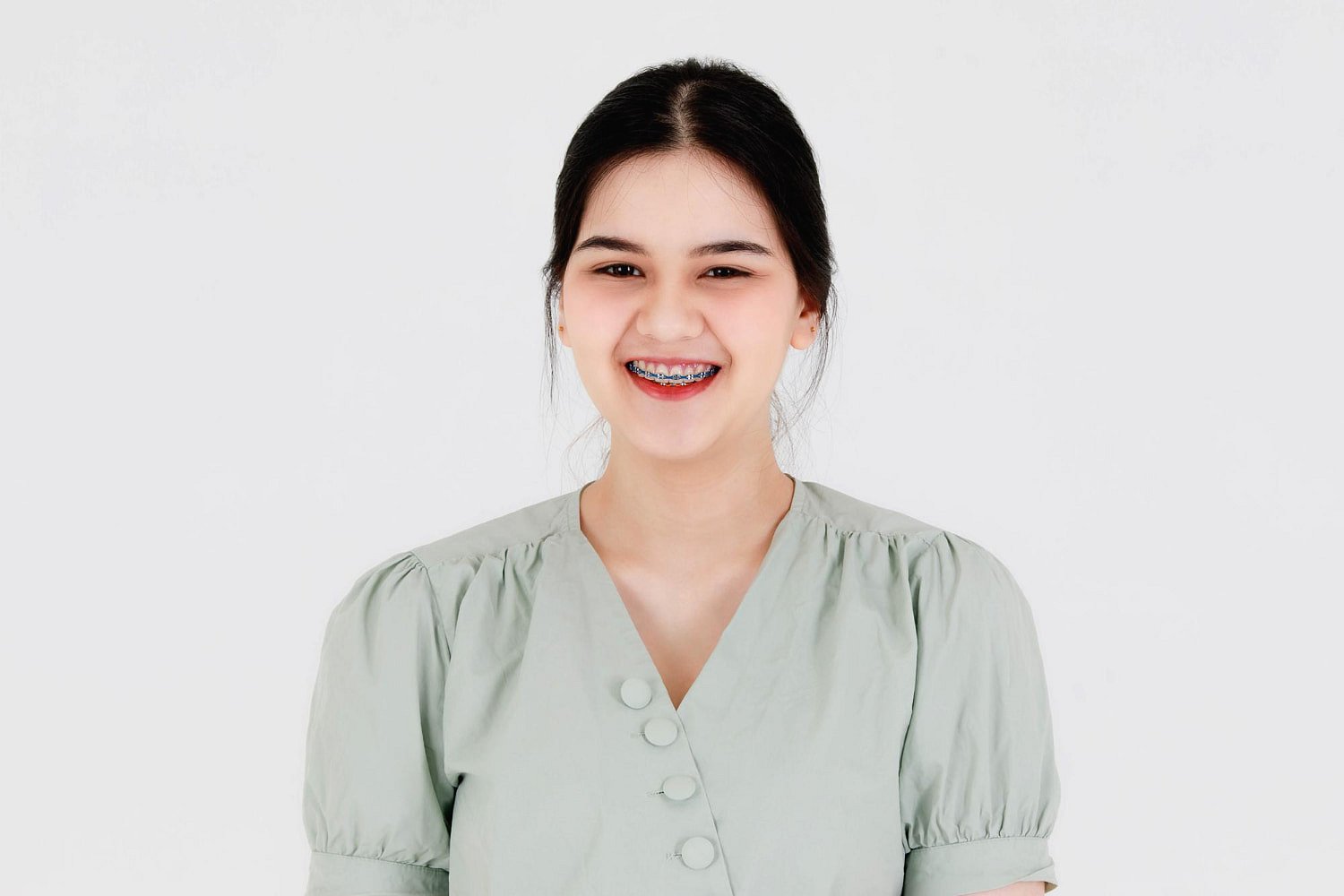 A young woman with dark hair in a light green blouse smiles at the camera, showing her braces, while considering Invisalign vs Braces at Andros Orthodontics in Pasco, WA.