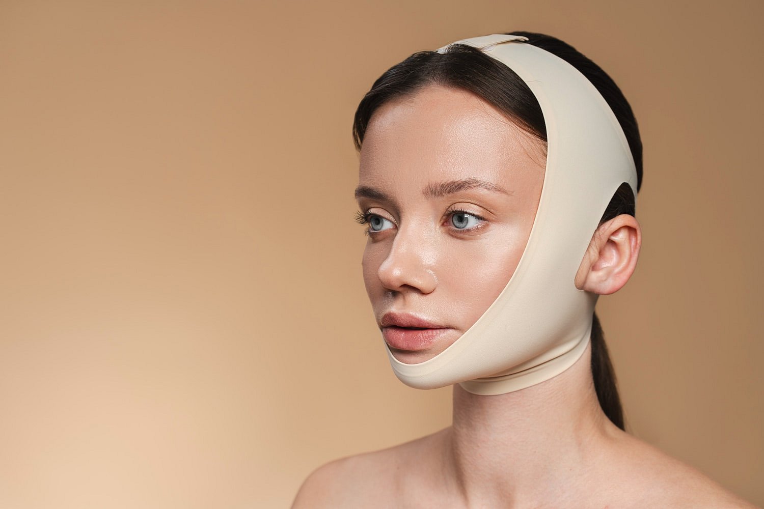 A woman in a beige compression facial bandage, typically worn post-surgery or for cosmetic recovery after Jaw Surgery, is shown against a plain beige backdrop at Andros Orthodontics in Pasco, WA.
