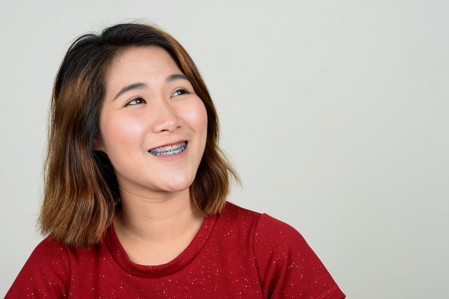 A young woman with shoulder-length hair, dressed in a red top and blue adult braces, smiles while looking to the side against a plain light background at Andros Orthodontics in Pasco, WA.