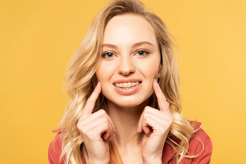 A young blonde woman smiles and points to her clear braces against a plain yellow background, showcasing the results of Andros Orthodontics in Pasco, WA.