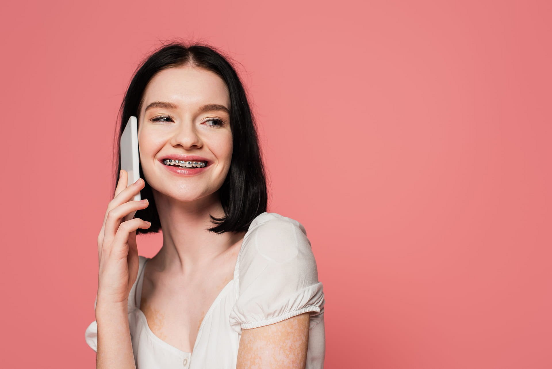 A young woman with dark hair and braces confidently smiles while holding a smartphone to her ear, highlighting the positive results of adult orthodontics at Andros Orthodontics in Pasco, WA, against a simple pink background.