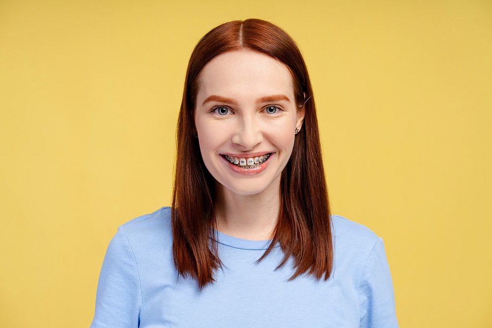 A young woman with straight red hair and metal braces smiles at the camera in front of a plain yellow background, representing Andros Orthodontics in Pasco, WA.