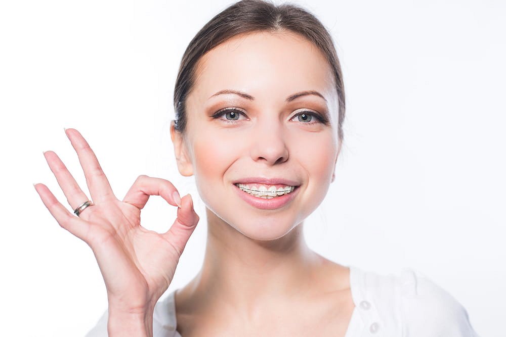 A young woman wearing clear braces smiles and gives an "OK" hand gesture, featured by Andros Orthodontics in Pasco, WA against a white background.