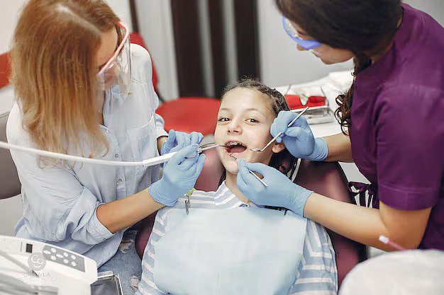 A young girl receives a dental checkup at Andros Orthodontics in Pasco, WA, as two dentists wearing gloves and protective eyewear discuss potential orthodontic appliances for her treatment.