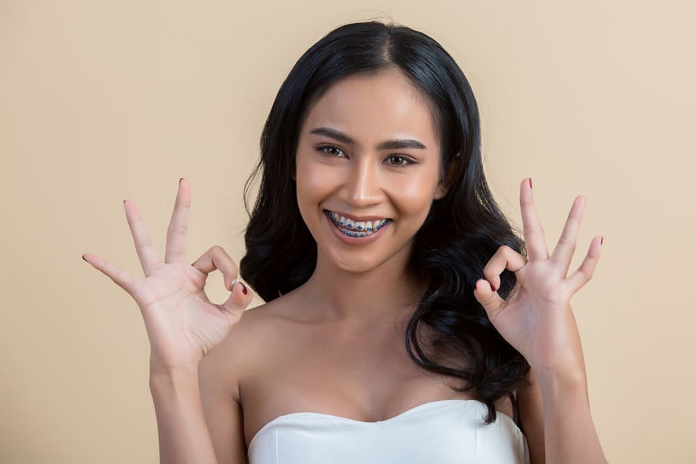 A young woman with long dark hair and Traditional Metal Braces, treated at Andros Orthodontics in Pasco, WA, smiles and flashes an "OK" gesture with both hands against a beige background.