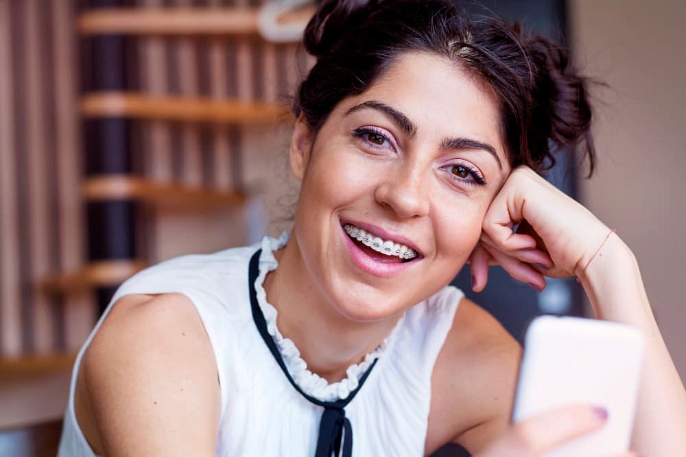 A young woman with adult braces at Andros Orthodontics in Pasco, WA smiles at the camera, holding her smartphone indoors with her elbow on a table.